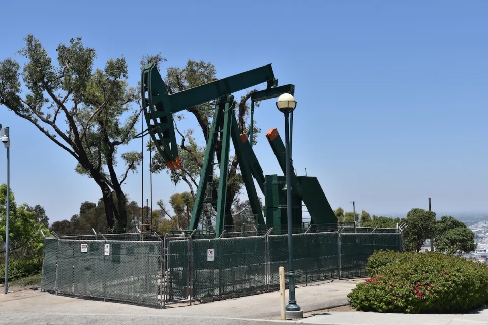 Green oil well pump jack surrounded by fence on a hill overlooking neighborhood.